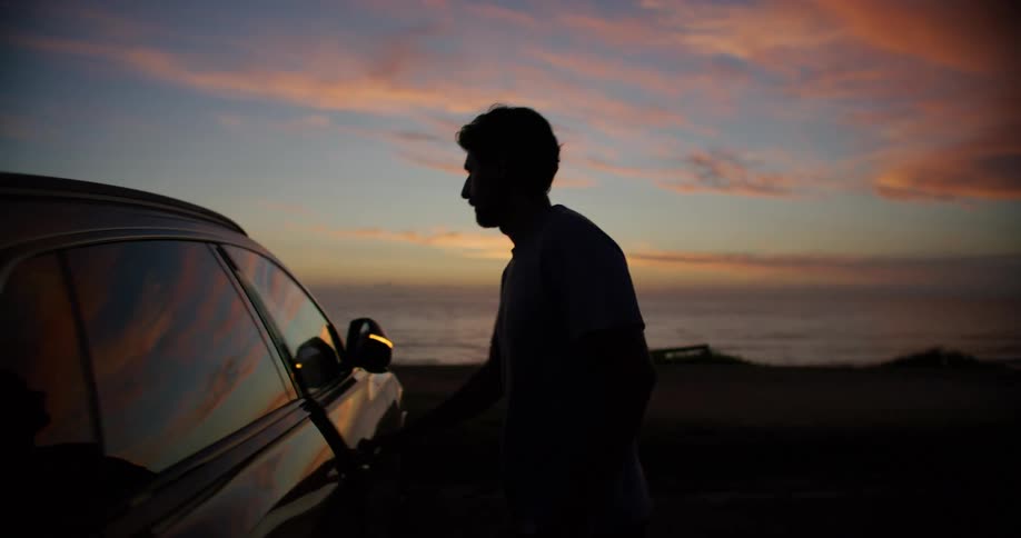 A silhouetted man approaches a parked car and opens the door against the backdrop of a vibrant sunset over the ocean