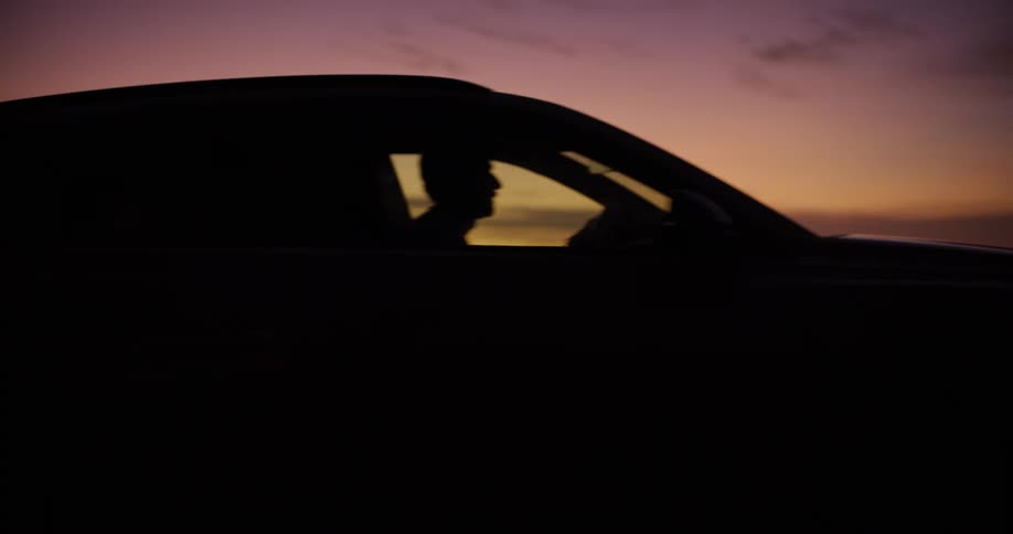 The silhouette of a vehicle and its driver against a vibrant orange and purple sunset sky with the car moving forward and exiting the frame followed by a wide shot revealing a blurred dark landscape with an ocean horizon and beach setting