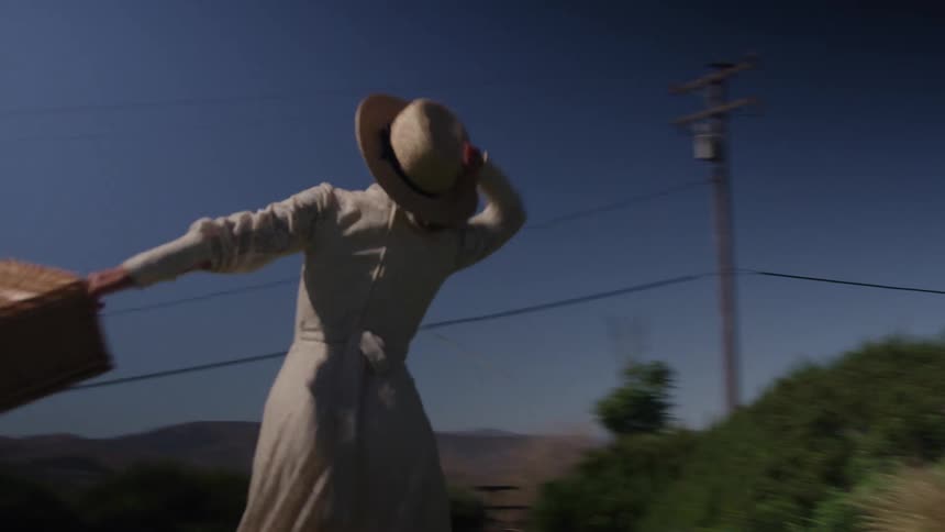 A medium shot of a woman in a long white dress and straw hat spinning outside while holding a wicker basket on a bright sunny day