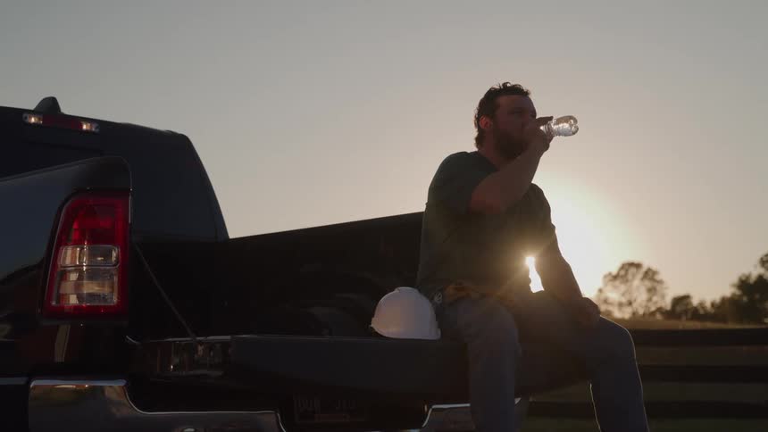 A man who appears to be a construction or manual worker is sitting on the open tailgate of a dark pickup truck silhouetted against a setting or rising sun he is seen taking a drink from a plastic water bottle with a white hard hat resting beside him