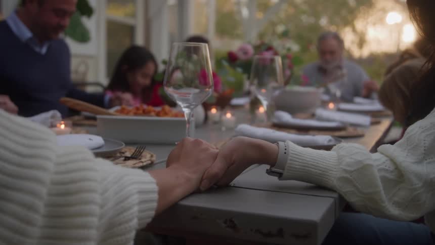 Family holding hands while praying together as they are seated at an outdoor dining table for a meal