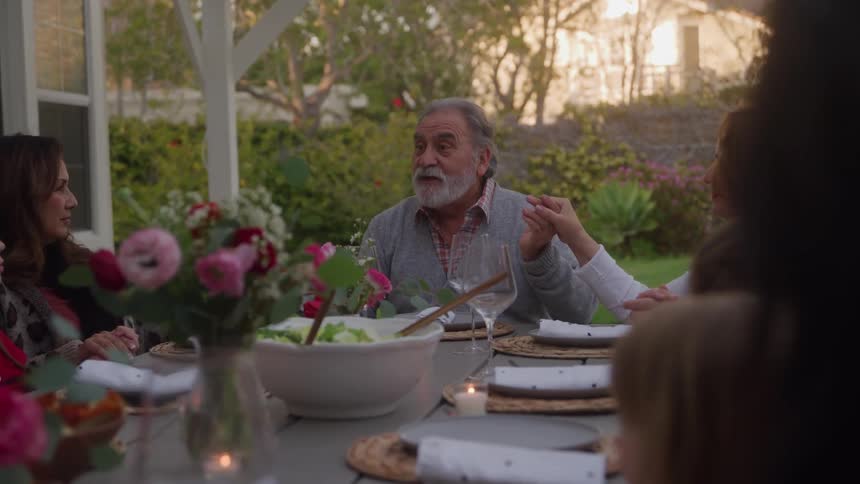 An older man with a beard and a serious expression looking forward while sitting at an outdoor dinner table with family or while holding a womans hand