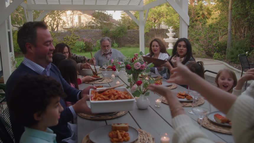 A large family shares a meal at an outdoor table under a white pergola as a man in a blue sweater accepts a large serving dish of pasta from a woman