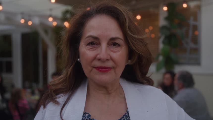 A medium closeup portrait captures an elegant older woman smiling warmly at the camera standing in a backyard during an outdoor family dinner party with blurred guests and string lights glowing behind her