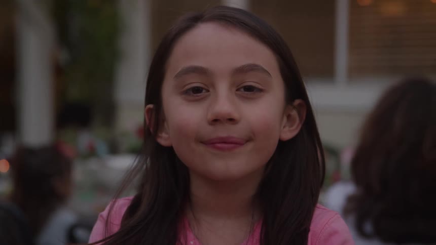 A closeup portrait of a young girl with long dark hair captures her engaging directly with the camera exhibiting a subtle sweet smile with people in the blurry background