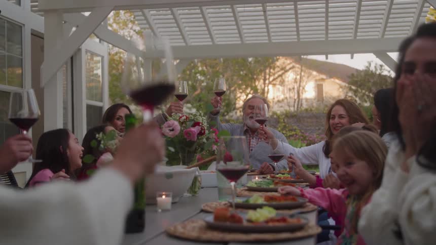 A multigenerational family and friends are gathered around an outdoor dining table under a white pergola during a dinner party or celebrationa smiling bearded older man raises his glass of red wine high for a toast which is mirrored by several o