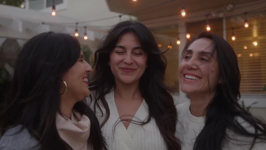Three women with long dark hair wearing lightcolored sweaters are captured in a closeup as they share smiles laughter and a warm embrace clearly enjoying each others company in a casual outdoor patio setting decorated with string lights
