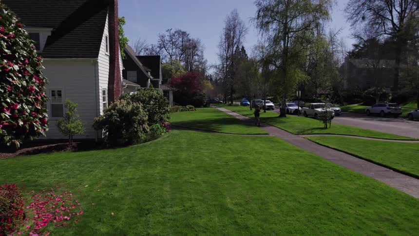 A long shot captures a tranquil suburban neighborhood street scene on a bright sunny day a person in athletic wear is jogging away from the camera on a sidewalk next to a lush wellmaintained green lawn