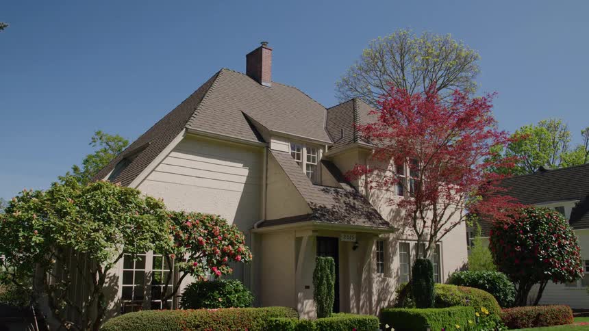 A wide shot of a traditional stucco house with a dark roof and a prominent chimney surrounded by neatly manicured green lawn hedges and flowering trees under a bright blue sky