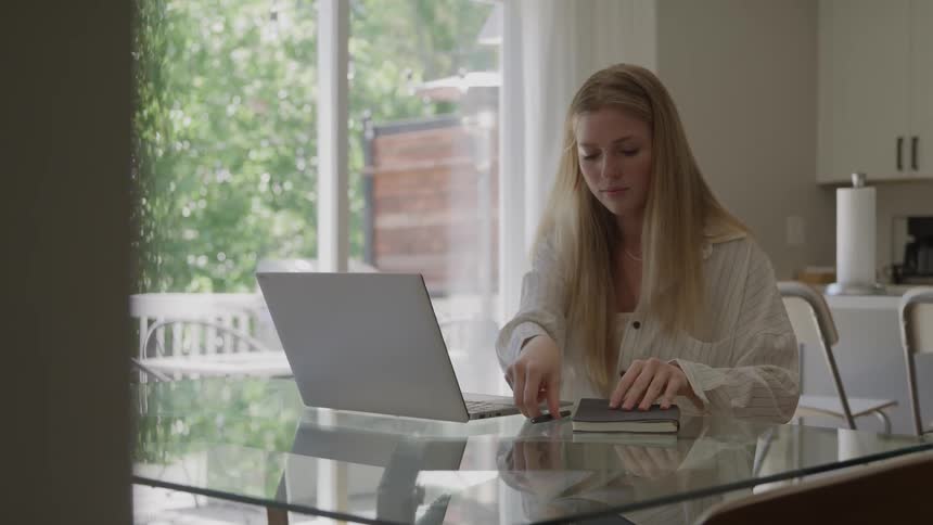 A woman walks to a table and sits down then opens her laptop and writes in a book
