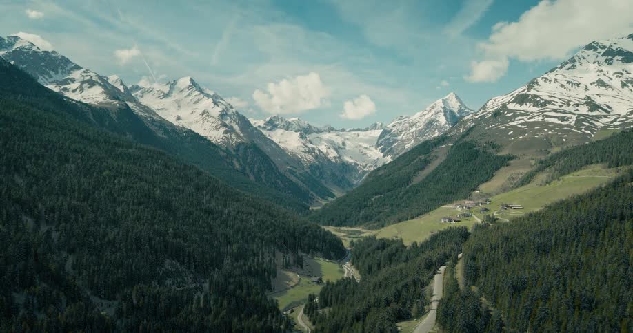 An aerial view of a mountain valley with snow capped peaks and a small village by a winding road