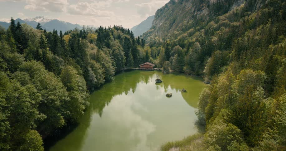 A secluded wooden cabin sits on the edge of a green lake surrounded by dense forest and towering mountains