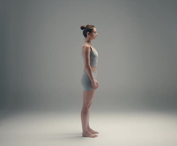 A woman in athletic attire stands barefoot in a neutral straight profile pose against a gray softly lit studio background with her arms relaxed at her sides