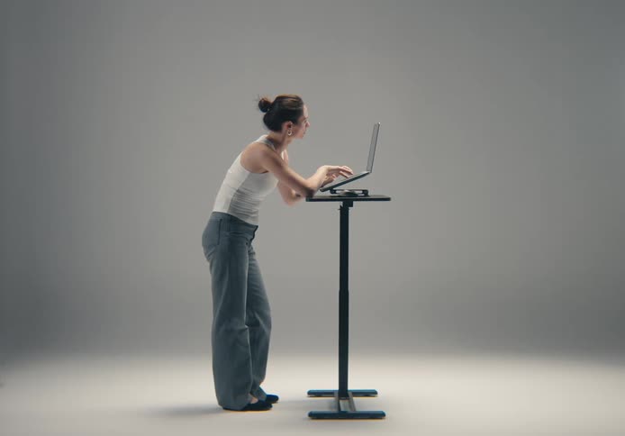 A woman in casual attire stands in profile view leaning slightly over a laptop placed on a tall slender standing desk while working intently in a bright neutral studio environment