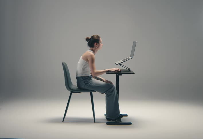 A focused woman in a white tank top and jeans is sitting while working on a laptop at a small table against a grey background