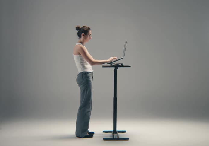 A focused woman in a white tank top and jeans is standing while working on a laptop at a small standing desk against a grey background