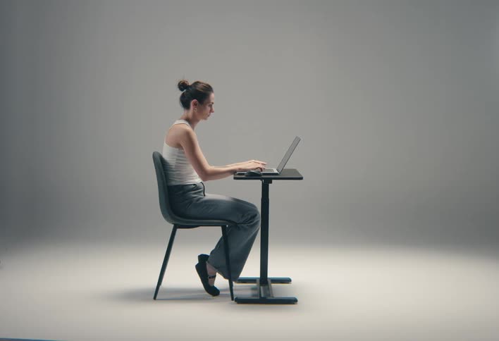 A young woman sits indoors behind a desk typing on her laptop