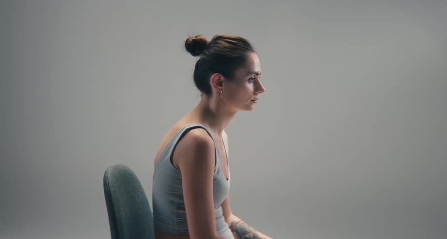 A woman sits in profile with a hunched posture in a plain softly lit studio