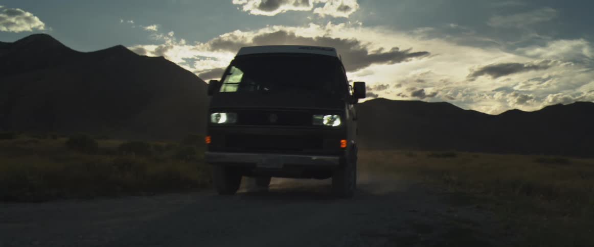 A van drives toward the camera on a dirt road and passes by backlit by a dramatic mountain sunset
