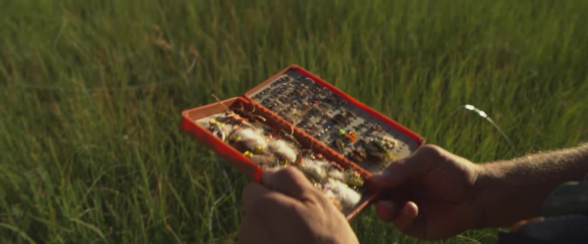 A man squats on a grassland at midday holding and opening a fly box