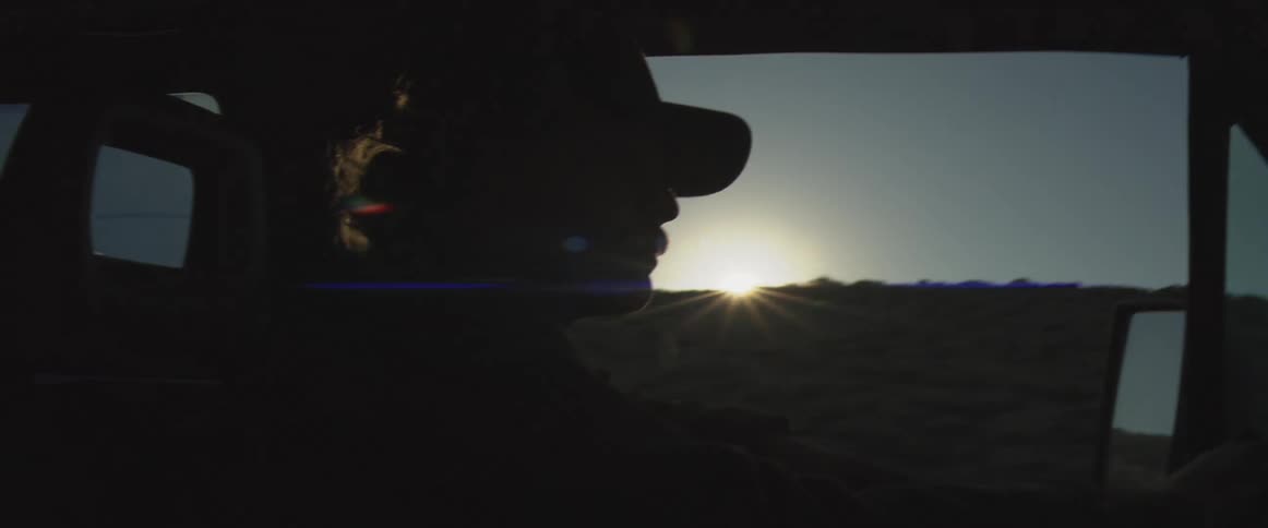 A dark silhouette of a person wearing a cap and driving a vehicle is visible from the side with the bright sun flaring over a dark rolling landscape outside the window