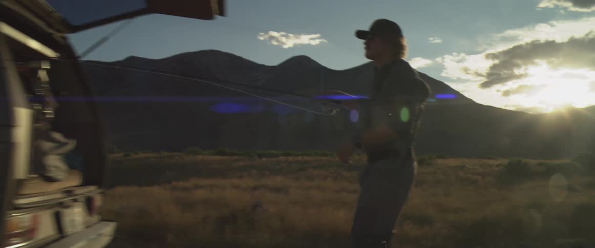 Man taking a fishing pole out of the back of his truck as the sun shines over mountains in the distance