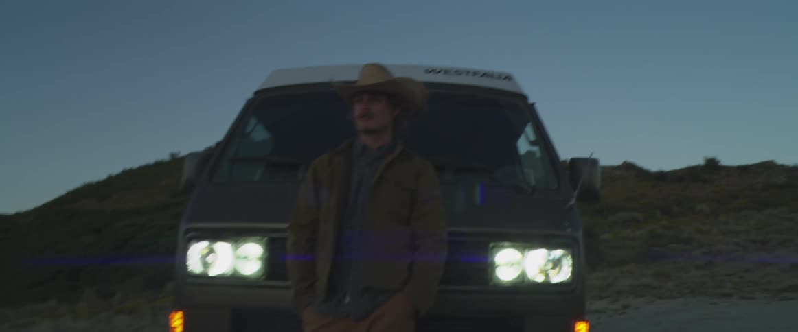 A man wearing a cowboy hat jacket and casual pants is standing in front of a volkswagen westfalia vanit is dusk or twilight and the vans headlights are on illuminating the man against the dark backdrop of a hillside