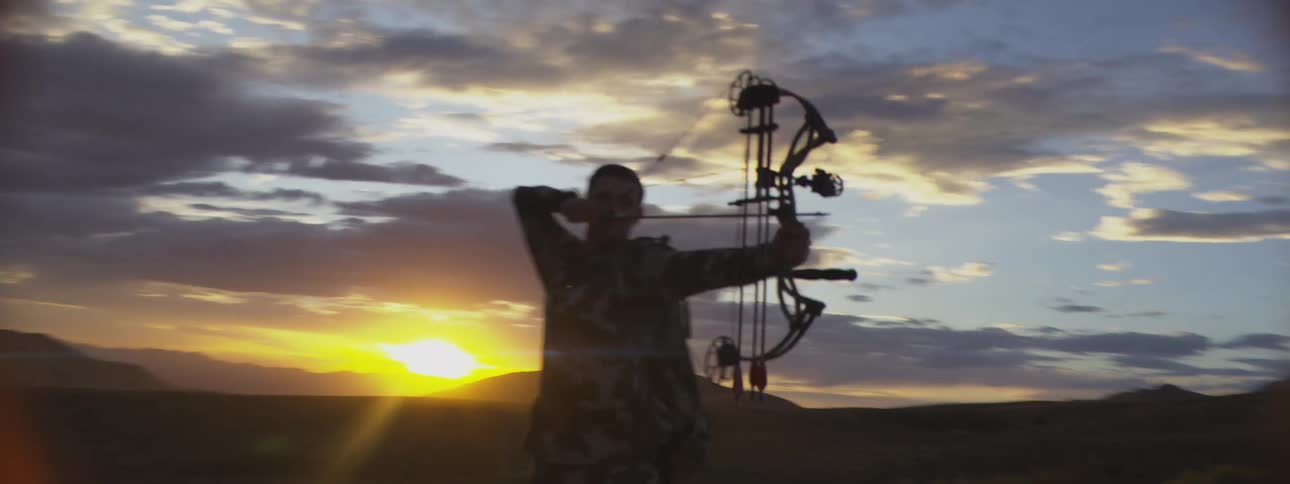 Man in camouflage gear drawing a compound bow in front of the sunset