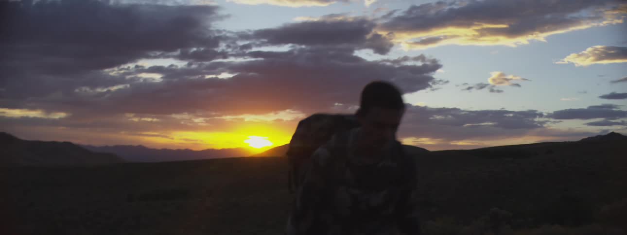 A wide shot captures a person wearing camouflage and carrying a pack walking across a ridge at sunset with the bright sun low on the horizon behind dark mountains and a colorful cloudy sky