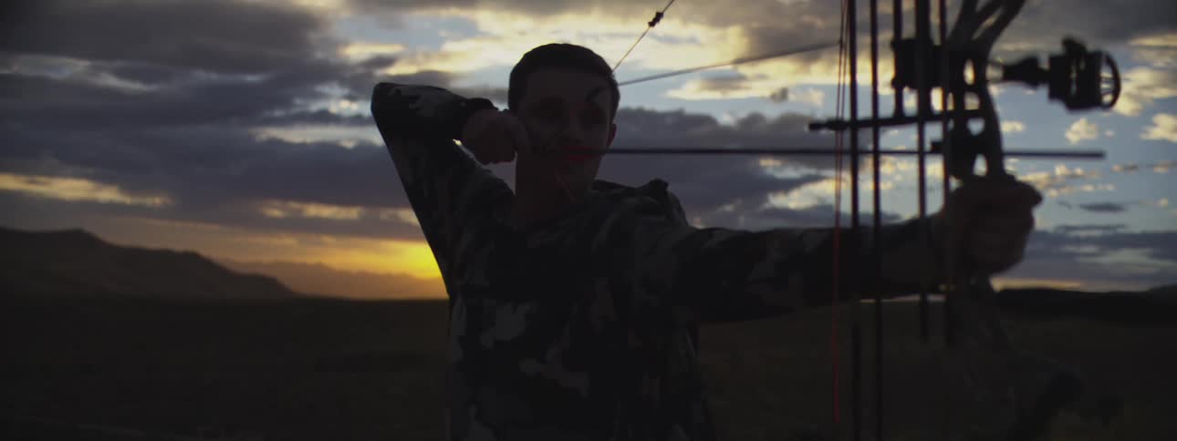 Man aims a compound bow and shoots at dusk in an open area