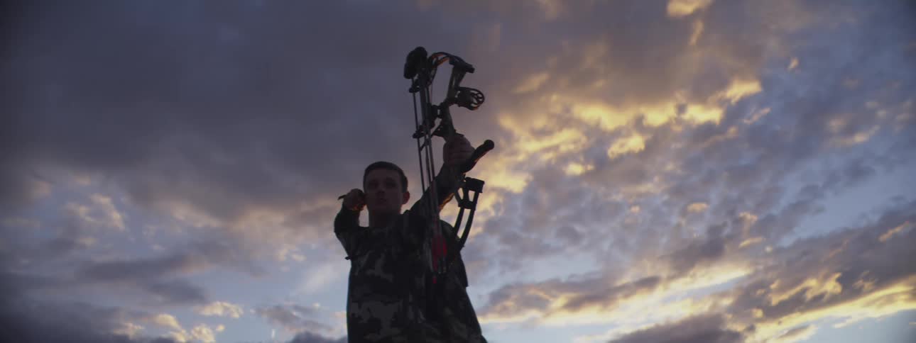 Man in camouflage gear drawing a compound bow under a cloudy sunset sky