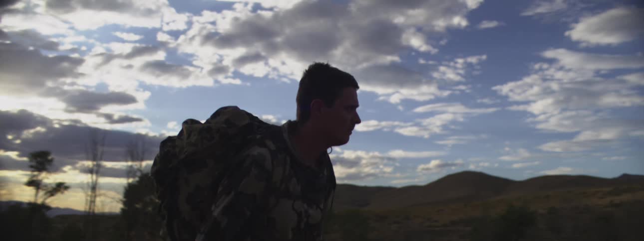 Man in camouflage gear walking with a backpack at sunrise among hills