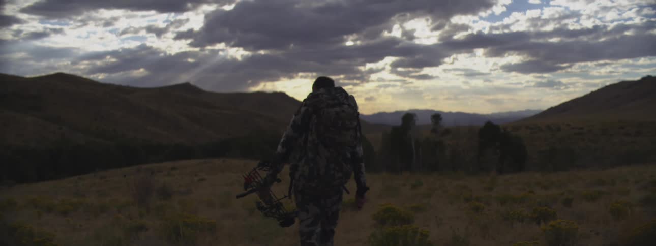 A rear shot shows a hunter wearing camouflage and a large backpack carrying a compound bow while walking through a vast hilly landscape at sunset under dramatic cloudy skies