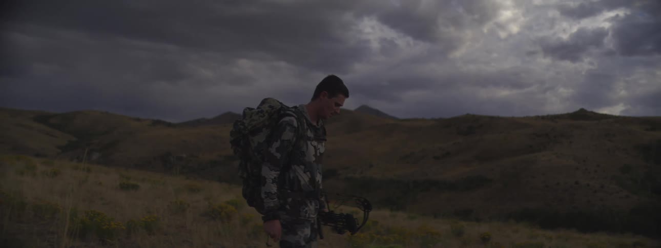 Man in camouflage gear walking with a backpack and a bow in a mountainous area