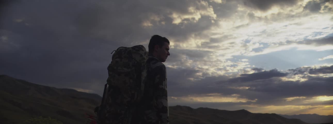 A camouflaged hunter holding a compound bow stands silhouetted against a dramatic sunset while overlooking a mountain valley