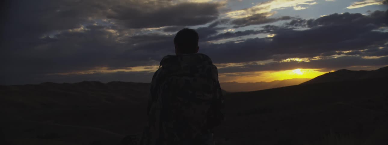 A silhouetted hunter stands overlooking a mountain valley during a dramatic sunset