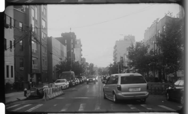 This is a short black and white clip of a city street filmed from the perspective of a moving vehiclethe scene features cars driving down a street lined with parked vehicles and multistory apartment buildings giving the impression of an older den