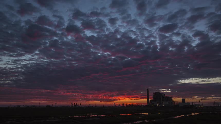 A time lapse of a vibrant sunset over a flat landscape with a large industrial plant silhouetted on the right and clouds changing from golden to dark blue and red