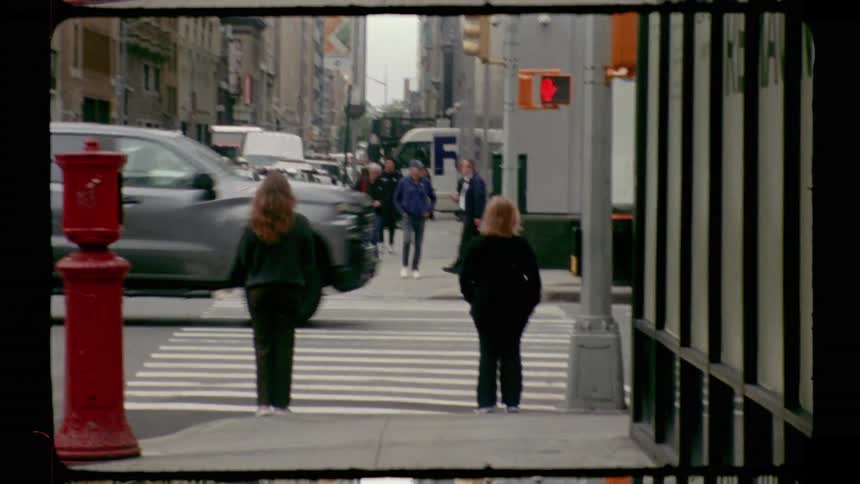 A vintage style film shot captures a woman waiting to cross a busy city intersection in new york city while people cross the street and traffic passes with tall buildings visible in the background