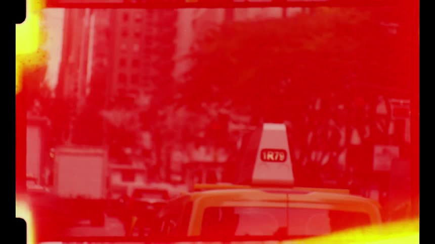 Yellow taxi and a line of vehicles moving along an avenue surrounded by tall buildings and trees