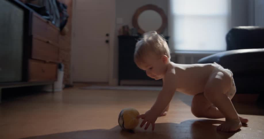 Diapered baby squatting on a wooden floor while watching a rolling ball