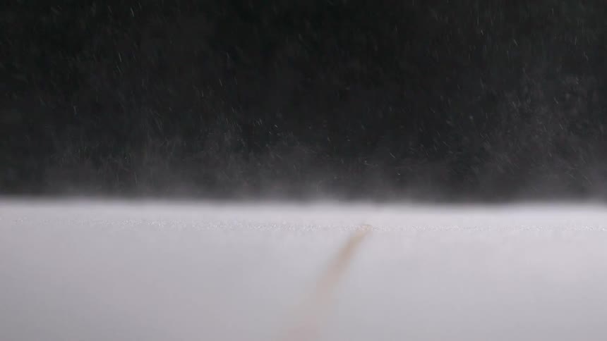 A close macro shot shows a fine stream of brown powder falling onto a white surface and creating a small pile before the rest is swept away by a puff of air
