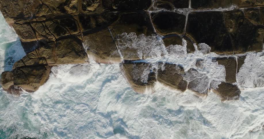Strong waves crashing against an irregular blocklike rock formation seen from above