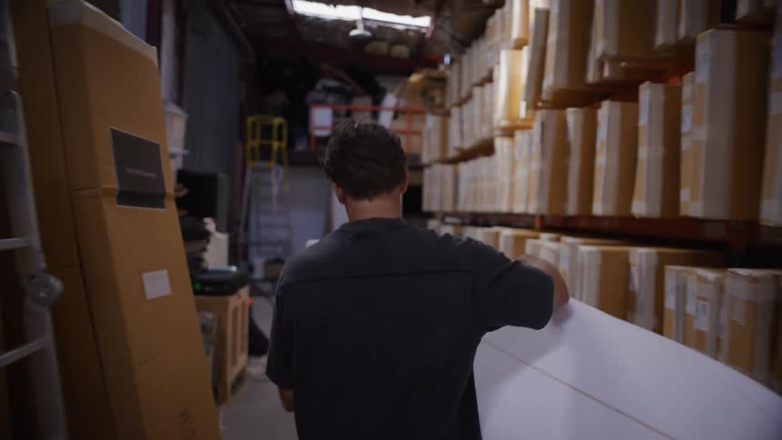 A man walking through a warehouse carrying a surfboard