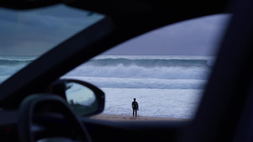 Viewed through a car window a lone figure stands on the beach facing large crashing ocean waves