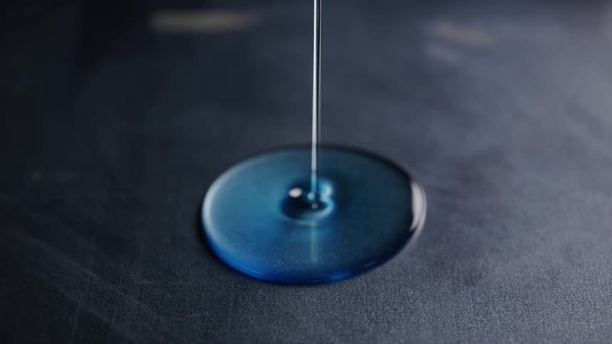 A macro close view shows a thin stream of viscous bright blue liquid slowly pouring onto a dark dusty surface spreading into a perfectly round puddle