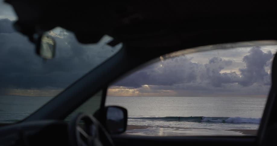 Car interior framing an ocean view with surfers under a cloudy sky