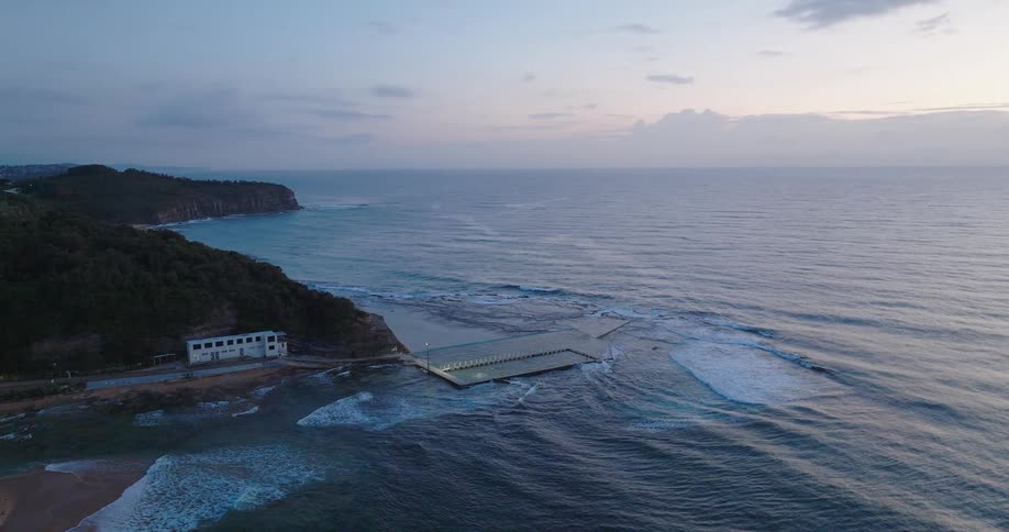 Waves from the ocean crash against the shoreline at sunset with a building standing nearby along the coast bathed in the warm glow of fading light