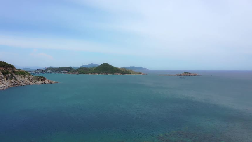 A wide deep blue ocean bay surrounded by green rocky hills and a few smaller islands with a distant village nestled along the left side of the rugged coastline