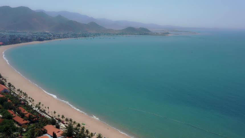 An aerial view shows a long curved sandy beach and coastal town with resorts facing a large bay of turquoise water with distant mountains under a hazy blue sky